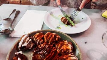 Woman cutting seasoned potato on her plate using knife and fork. Shared dish with potatoes covered in smoked paprika, mayonnaise, and ketchup is placed in center of table. Cozy restaurant setting video