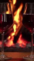 Two glasses of red wine in front of a fireplace, A romantic setting with two glasses of red wine placed on a wooden surface in front of a warm, glowing fireplace. Vertical footage video