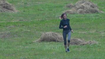 Female runner in sportswear - tracksuit jacket, cropped leggings and sneakers, running across field grass during sports training, summer physical activity. Full length front view, real time. Zoom out video