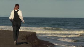 Full length view of unrecognizable woman running along black sandy beach during summer holiday. Model is dressed in sweater, denim jumpsuit and trainers. Real time, cinematic handheld shot, zoom in video