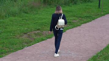 High angle rear view of an unrecognizable woman walking on footpath in public park in summertime. Model wears blue jacket, jeans and carries small backpack on her back. Handheld tracking shot, zoom in video