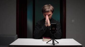 Priest praying with rosary in front of smartphone on tripod with bible on table video