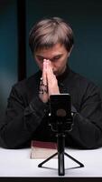 vertical Priest praying with rosary in front of smartphone on tripod with bible on table video
