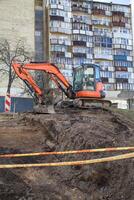A bulldozer digging a foundation pit and operating at a construction site among residential buildings, behind a safety tape photo