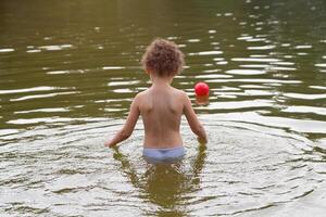 A curly child plays with a ball in the lake in the summer photo