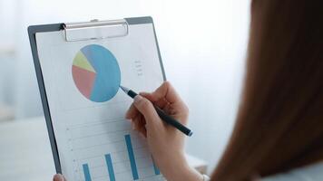 A young businesswoman focuses on chart analysis while working in a bright, open office. She holds a clipboard and uses a pen to make notes, showcasing her corporate lifestyle and dedication. video