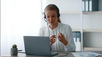 A young businesswoman is participating in a virtual meeting from her stylish office workspace. She is focused and actively discussing, wearing a headset and gesturing with her hands. video