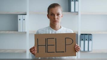 In a modern corporate setting, a young woman holds a large cardboard sign that reads 'HELP'. Her serious expression conveys the need for assistance in her professional environment. video