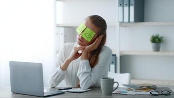 A young woman in a corporate office setting sits at a desk, using sticky notes to cover her eyes while taking a moment of relaxation. She appears thoughtful and enjoys the calm atmosphere. video