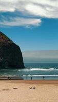 Giant Haystack Rock formation on a sandy beach with visitors. A towering rock formation stands tall on a sandy beach as visitors walk along the shore. video