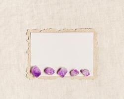 Square invitation card mock up, blank greeting card and gemstone crystal, healing stone violet amethyst on beige table. Torn paper. Minimal business brand template. Flat lay, top view photo