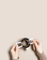 Woman hand holding cup and make black coffee drink in cup, drip coffee bag with ground coffee brewed in cup, paper bag drip coffee filter, beige background, top view, minimal flat lay photo
