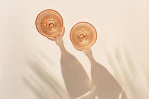Two wine glasses from peach color glass with palm leaf shadows from sun, trendy goblets style wine glasses with white wine on beige background. Pastel colors, monochrome, top view glassware photo