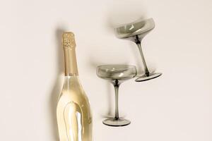 Champagne or sparkling wine bottle, champagne glasses from tinted grey glass on beige background. Festive drink minimal concept. Modern wine glasses, dark colored glass. Creative top view, flat lay photo