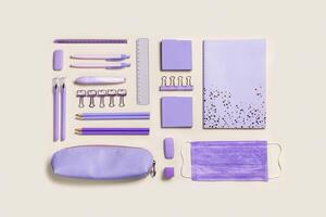 School stationery flat lay violet color. Back to school new normal concept, notebook, note, pen, pencil, clips, medical mask on beige background. Office equipment pattern, top view photo