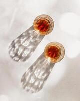 two Whiskey Glasses with Shadows and sunlight Reflection on beige background, minimal aesthetic top view summer still life, refraction throug crystal shine cups, caustics effect, vertical photo
