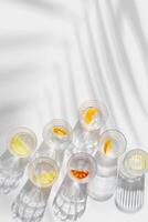 Fruit Infused Water in transparent Glasses, palm leaf Shadow at sunlight on White background with caustics effect. Aesthetic summer fresh drink with slices of lemon and orange in glass cups, top view photo