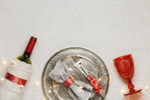 Christmas rustic table setting, red wine glass, plate, knife, fork, wine bottle, decorated green fir tree on sacking tablecloth. New Year dinner with xmas decor, serving festive table photo