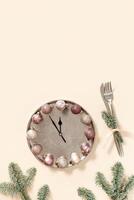 Winter festive table setting concept, Christmas plate with clock hands show twelve or midnight, sparkle balls beige pink color, knife, fork, fir tree branches. New Year holiday food concept photo