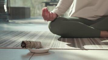 Sunlight illuminates a woman practicing yoga and meditation with her hands in a mudra position, while a smoldering white sage bundle rests on a small plate in front of her video