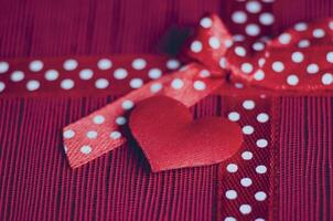 Side view of red tiny heart and gift ribbon with white polka dots tied into a bow, placed on a textured red and pink striped background photo