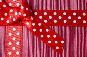 Close up of a red gift ribbon with white polka dots tied into a bow, placed on a textured red and pink striped background photo