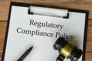 Regulatory Compliance Policy text on clipboard surrounding with magnifying glass and gavel. Compliance concept photo