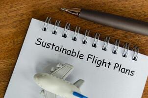 Sustainable Flight Plans text on white notepad with small airplane and pen background. Environmental awareness photo