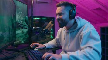 Smiling happy Caucasian addicted male gamer guy man streamer play at computer internet virtual gaming at home web battle online playing game at dark evening with microphone and headset laughing video