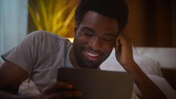 Happy cheerful smiling African American ethnic man male guy client user holding digital tablet using device watching online browsing internet shopping lying on bed at home bedroom late evening video