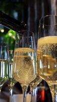 elegante champanhe ser derramado para dentro flautas às uma sofisticado evento, detalhado fechar-se do espumante vinho o preenchimento óculos com uma exuberante verde fundo e festivo atmosfera. vertical video