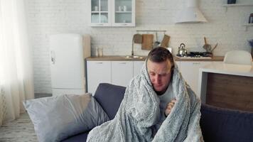 Man suffering from flu. Young sick man sitting on a sofa warming with blanket in the kitchen. Miserable man having a headache. Covid-19. Isolation at home. video