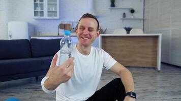 Smiling young man with a bottle of water at home. Young man proposing fresh water after the workout and looking on camera indoors. video