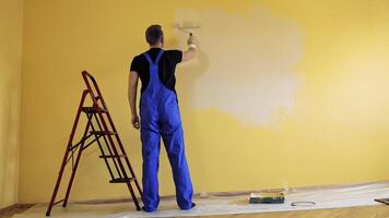 Room decoration with new color. Professional worker in blue overalls painting wall with a paint roller in empty room. Rear view. video