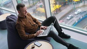 Young freelancer uses laptop. Handsome guy is sitting by the window with city view background and working on business with a wireless computer. video