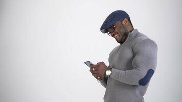 Stylish guy with black skin uses smartphone. Profile view of african man in cap and glasses typing messages on his mobile phone and smiling. video