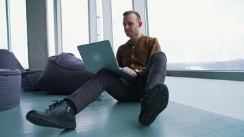 Young freelancer working on a laptop. Man in shirt and trousers sitting on a floor and using a gadget to work in office. Panoramic windows view. video