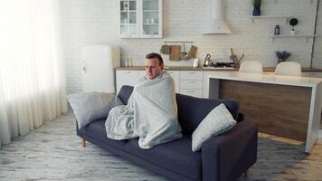 Man having fever. Young man feeling freezy and covering with warm blanket while sitting on a couch at home. Sick man wrapped with blanket during covid outbreak. video