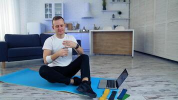 Man drinks water after daily workout at home. Guy is sitting on a fitness mat next to the laptop and holding a bottle of water. Fitness indoors. Sport during quarantine. video
