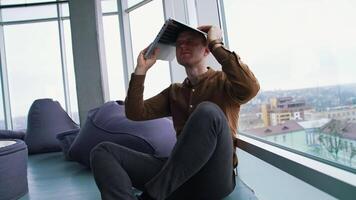 Tired businessman with a laptop on his head. Man is sitting in the office by the large window and holding a laptop over the head. Problems in business. video