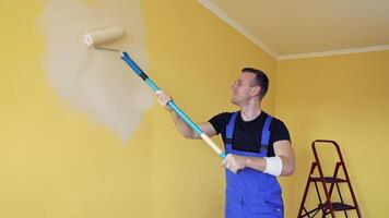 Room improvement with a new color. Professional worker in blue overalls painting wall with a paint roller on a long handle on. Craftsman working. video