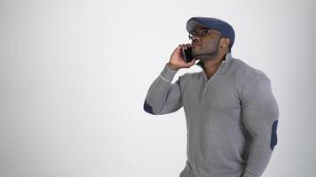 Black guy talks the phone. Afro american man in gray sweater and stylish cap leading a business conversation on white studio background. Slow motion. video