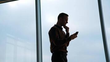 Young man on large window background. Businessman with a phone standing in office and looking through the window in the evening. Rear view. video