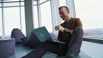 Smiling young man in modern office. Happy freelancer sitting on a floor and working on a laptop on window city view background. video