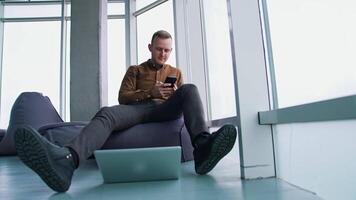Young man uses the mobile phone. Businessman is sitting in ottoman with a phone on large windows background. Laptop on the floor in office. View from below. video