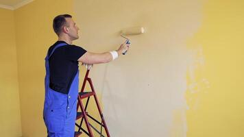 Man in blue overalls paints room. Worker paints the walls in the room with a paint roller on a ladder. Room decoration process into new color. video