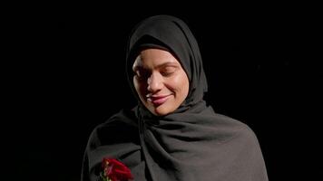 woman dressed in hijab holds red rose close her chest and smell it. Her expression reveals warmth and contentment as she stands in front of dark backdrop video
