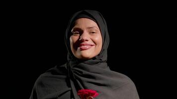 woman wearing black hijab stands confidently smiling gently while holding red rose and smell it. background is dark highlighting her joyful expression and vibrant flower video