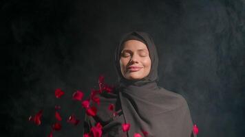 woman dressed in black hijab stands still her eyes closed in quiet space. Soft smoke surrounds her while red petals float gently above enhancing tranquil atmosphere video