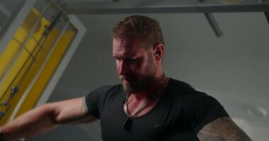 man wearing headphones engages in crossover hand raises at a gym showcasing his strength and concentration. His muscular build is highlighted by the close up perspective of the exercise video